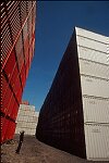 Shipping containers piled high in depots near ports in the U.S.A. illustrate America's trade deficit. Near Port Newark, New Jersey, container depots use special equipment to stack containers up to eight (8) high. Trucks drop off empty containers for storage or pick up empty ones for use. Depots resemble cities or monuments, blocking the skylines of Newark, N.J., and New York City.